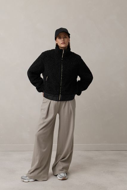 Person trägt schwarzen oversized Shearling-Jacken mit grauen weiten Hosen und Cap, steht lässig posierend.